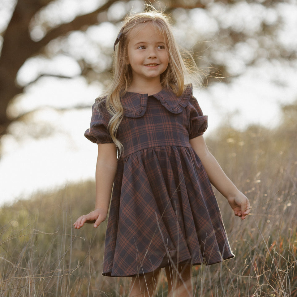 A young girl with long blonde hair stands in a field wearing a dark brown and red plaid dress with a ruffled Peter Pan collar.
