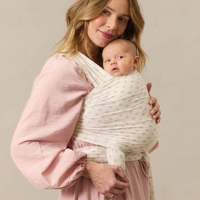 Woman in a pink dress holding a baby in a white wrap against a beige background
